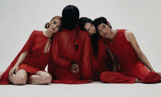 Four models wearing red outfits pose closely together on a seamless light gray studio background. Two women and two men are visible, styled in matching red garments with one figure facing away in a sheer red robe.