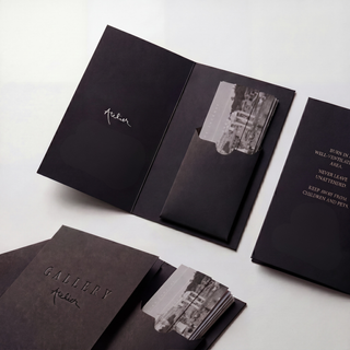 A set of black paper cardstock packaging pieces for Gallery Atelier, including an open tri-fold insert with printed text and a photo, a closed matching folder, and a stack of paper sticks, arranged on a light gray background.